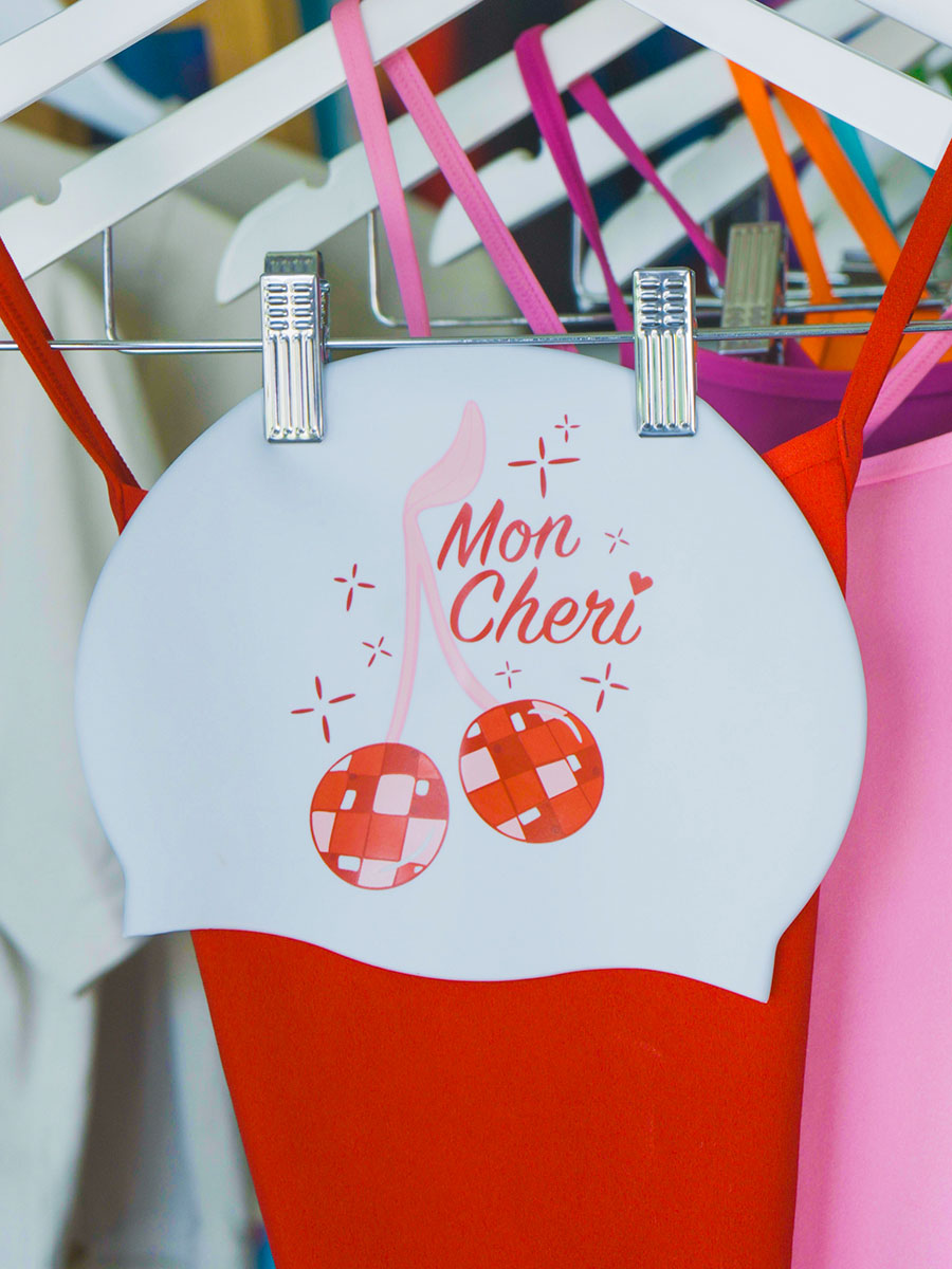 White, pink and red JAYD swim cap featuring cherry disco balls, hanging next to a red matching JAYD one piece swimsuit.