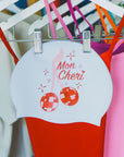 White, pink and red JAYD swim cap featuring cherry disco balls, hanging next to a red matching JAYD one piece swimsuit.