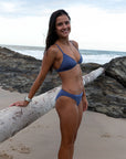 JAYD Swimwear model Ana wearing the Storm blue Kiama top and Cali bottoms, leaning against a log at the beach with waves breaking in the background.