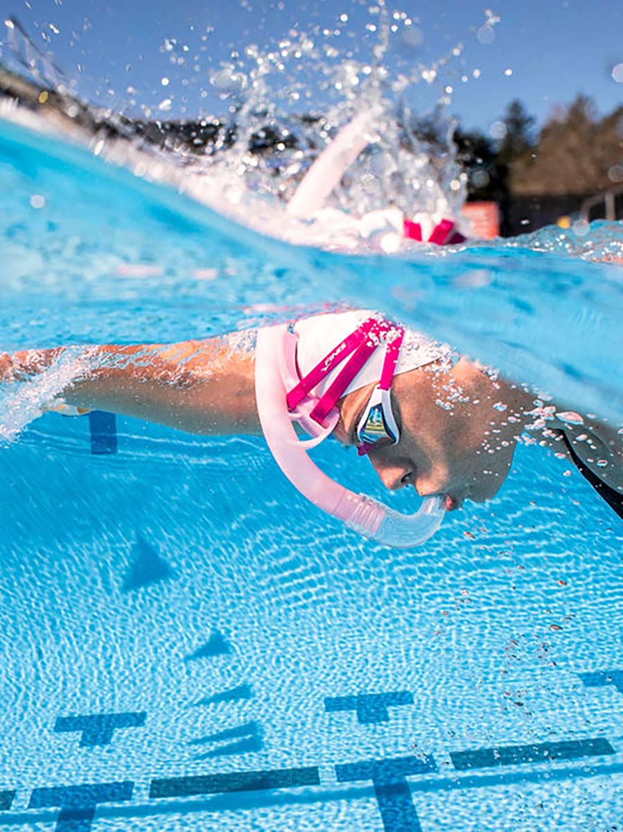 Underwater close-up of a swimmer wearing a pink FINIS Stability Snorkel snorkel and goggles in a blue swimming pool, with water splashing above the surface.