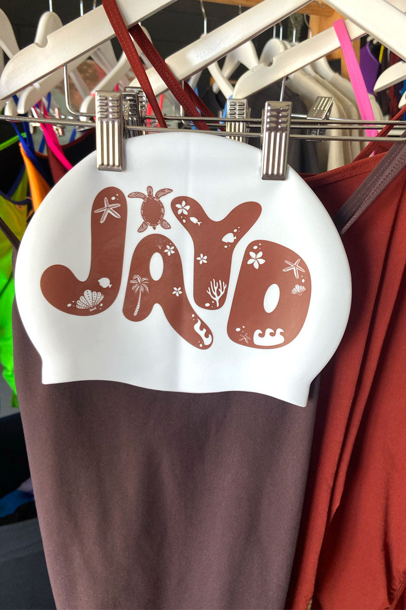 White swim cap with cacao 'JAYD' printed on it, in bold bubble letters, with tropical palm tree & turtle icons hanging on a rack with JAYD Swimwear cacao and cinnamon one pieces.