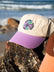 Salty Summer embroidery design on the cream JAYD Swimwear baseball cap with purple brim, sitting on a rock with the ocean in the distance.