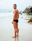 Athlete Wes wearing the JAYD Swimwear men's Ramshark black bottoms on a beach, looking over his shoulder at the camera.