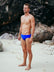 JAYD Swimwear model Wes wearing the cobalt blue Ramshark men's bottoms, standing on a sandy beach with rocky background.