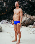 JAYD Swimwear model Wes wearing the cobalt blue Ramshark men's bottoms, standing on a sandy beach with rocky background.