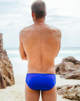 Male model wearing the JAYD Swimwear cobalt blue Ramshark men's swim bottoms, standing on a beach with rocks and ocean in the background
