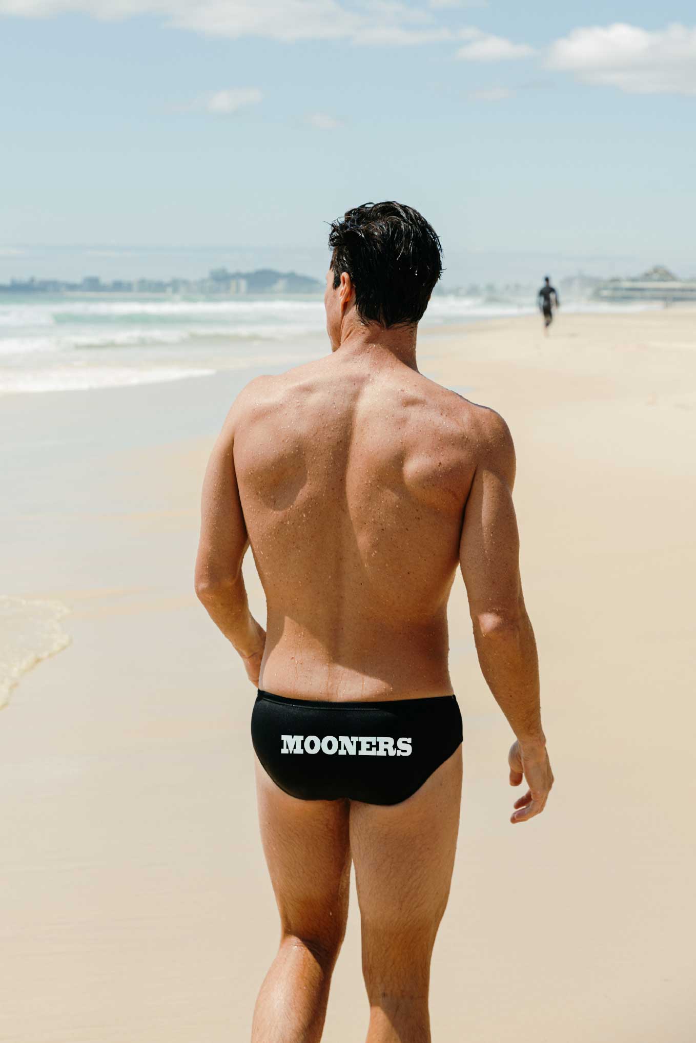 Man walking on a beach wearing black swim briefs with customised 'MOONERS' branding.