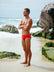 Athlete Wes wearing the JAYD Swimwear Ramshark mens bottoms in red, standing on a rocky beach staring into the distance.