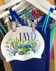 White swim cap design featuring JAYD logo & blue and green coral illustrations, paired with JAYD one piece blue swimsuits.