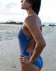 Side view of the storm blue JAYD Riva one piece swimsuit, with model Ana looking into the distance on a sandy beach
