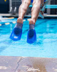 A close-up action shot of a person's legs and feet wearing bright blue FINIS Swim SlipFin flippers jumping into a swimming pool, with water splashing and the pool deck in the foreground.