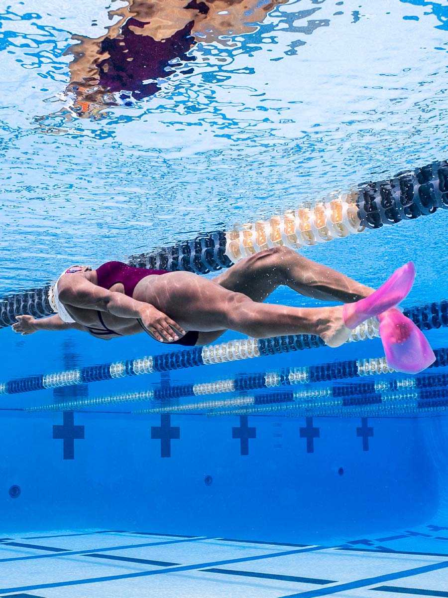A person underwater in a swimming pool, wearing a dark purple and black swimsuit with bright pink FINIS Swim SlipFin flippers. Lane ropes and blue pool water are visible in the background.