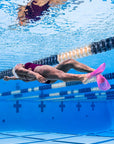 A person underwater in a swimming pool, wearing a dark purple and black swimsuit with bright pink FINIS Swim SlipFin flippers. Lane ropes and blue pool water are visible in the background.