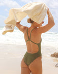 Back view of model Brodee Trinca wearing the JAYD wimwear Soraya One Piece in Olive with a towel blowing in the wind.