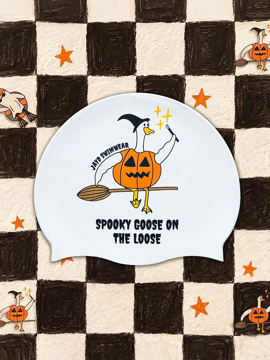 A white novelty swim cap featuring a cartoon illustration of a goose dressed as a witch riding a broomstick on a pumpkin, with the text 'JAYD SWIMWEAR' and 'SPOOKY GOOSE ON THE LOOSE'. The cap is displayed on a black and cream checkered background decorated with small orange stars and cartoon pumpkins.