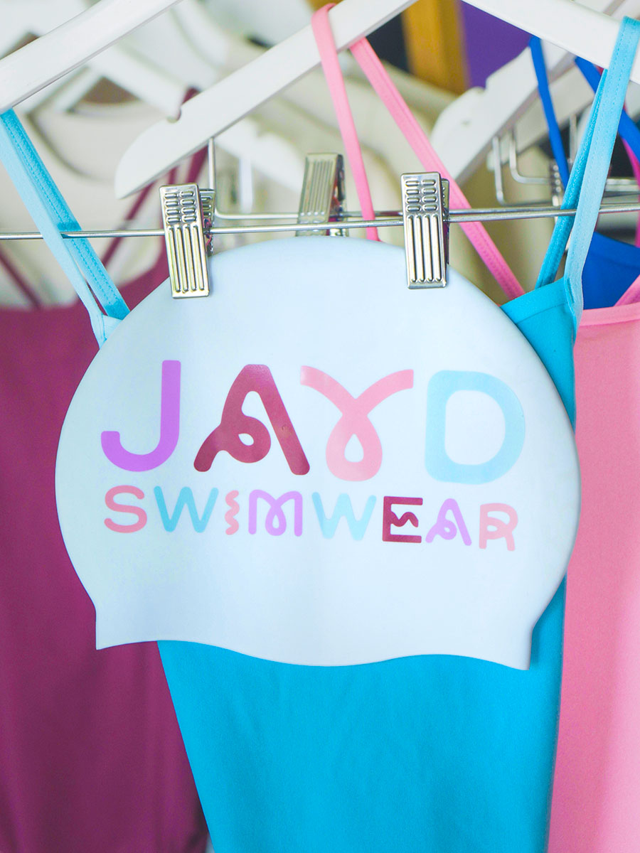 White swim cap with squiggly colourful 'JAYD Swimwear' branding on a rack with complimentary sangria, fairy floss and cancun one pieces.