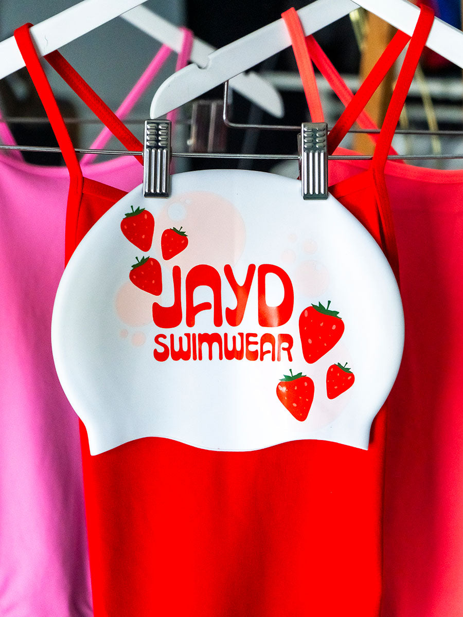 White swim cap with red JAYD Swimwear text and floating strawberries.