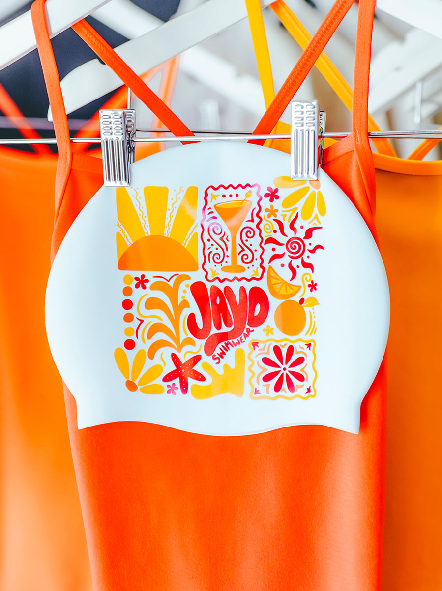 White JAYD Swim cap with colourful collage style illustrations hanging on a rack with swimsuits in the background