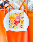 White JAYD Swim cap with colourful collage style illustrations hanging on a rack with swimsuits in the background