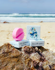 JAYD Swimwear pink circular surf wax arranged in a stack on top of a rock with the beach and ocean in the distance.