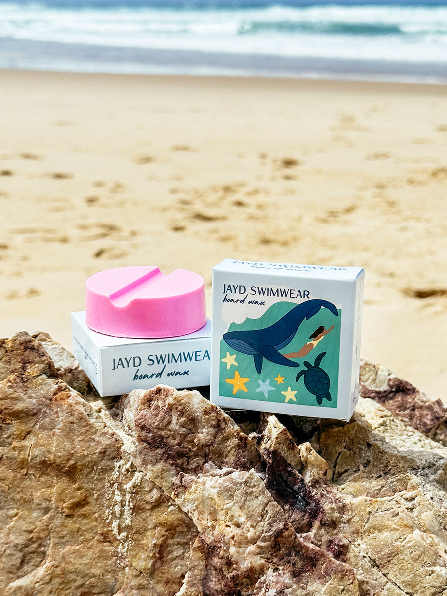 JAYD Swimwear board wax sitting on a rock at the back, showcasing packaging illustration of a woman swimming with a whale and turtle.