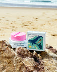 JAYD Swimwear board wax sitting on a rock at the back, showcasing packaging illustration of a woman swimming with a whale and turtle.