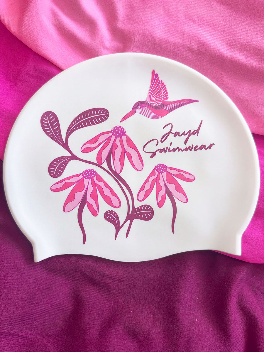 A white swim cap featuring a pink and magenta floral design with a hummingbird and the text 'Jayd Swimwear', displayed on a draped magenta fabric background.