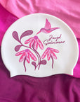 A white swim cap featuring a pink and magenta floral design with a hummingbird and the text 'Jayd Swimwear', displayed on a draped magenta fabric background.
