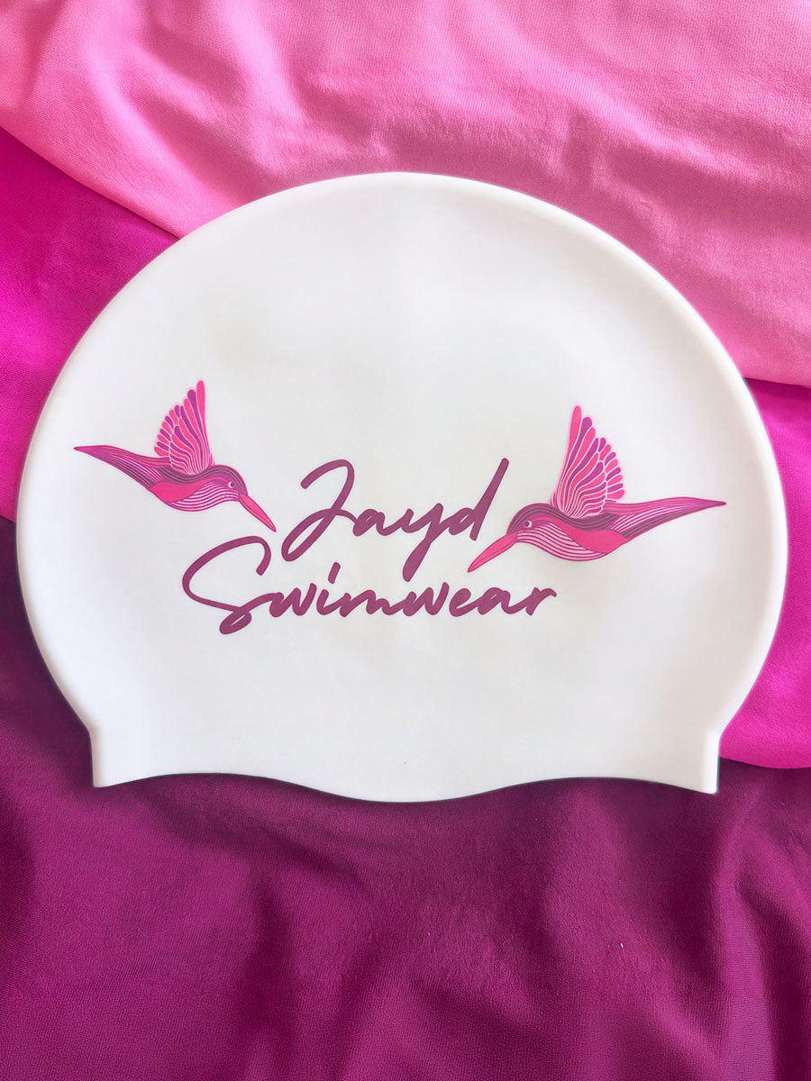 A white swim cap with pink printed graphics of two hummingbirds and the text 'Jayd Swimwear', displayed on a pink and magenta fabric background.