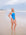 JAYD Swimwear model in a bright Cancun blue one-piece Tallow swimsuit on a sandy beach with the ocean and cloudy sky in the background.