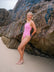 JAYD Swimwear model on a sandy beach leaning against a large rock wall, wearing the bright pink fairy floss Tallow one-piece swimsuit.