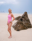 JAYD Swimwear model wearing the fairy floss pink Tallow one-piece swimsuit, stands on a sandy beach next to a large rock, with the ocean and cloudy sky in the background.