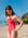 JAYD Swimwear model wearing the Tallow one piece swimsuit in Fiesta, standing on a sandy beach with the ocean and rocky coastline in the background under a clear blue sky.