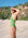 JAYD model Kiera wearing the moss green one-piece swimsuit stands on a sandy beach with rocky cliffs and greenery in the background, shielding her eyes from the sun.