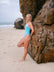 JAYD Swimwear model posing on a sandy beach next to a large rock, wearing the light blue Mykonos one-piece Tallow swimsuit.