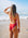 JAYD model standing on a beach looking out across the water, wearing the full bum coverage Tallow one piece swimsuit in bold ruby red.