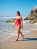 JAYD Swimwear model walking along he shoreline of a rocky beach, wearing the Tallow one piece swimsuit in ruby.