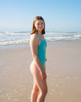 JAYD Swimwear model Amelia wearing the Tallow seamist one-piece swimsuit, standing on a sandy beach with the ocean and blue sky in the background.