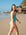 JAYD model Amelia woman wearing the seamist Tallow one-piece swimsuit, walking along a sandy beach with ocean waves and rocky cliffs in the background.