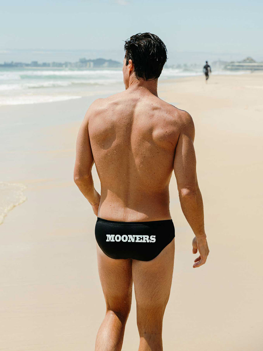 Man walking on a beach wearing black swim briefs with customised 'MOONERS' branding.