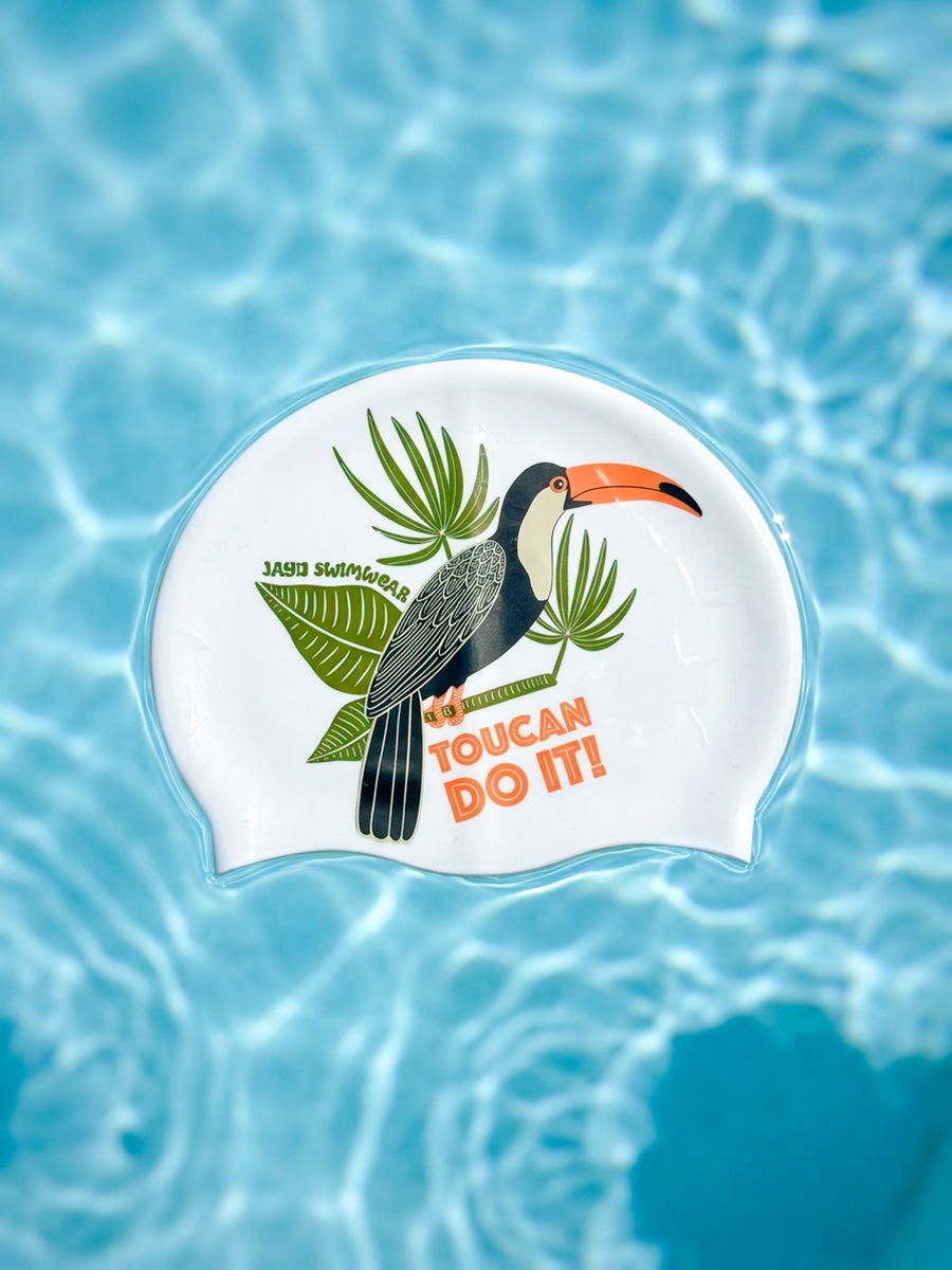 A white swim cap featuring a colorful toucan illustration and the text 'TOUCAN DO IT!' floating on clear blue pool water.