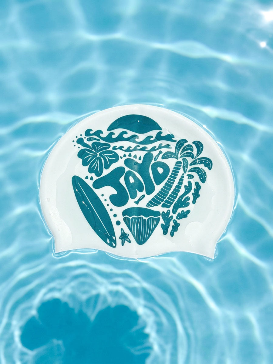 A white swim cap with a teal graphic design featuring the word 'JAYD' and tropical motifs like palm trees, a surfboard, and flowers, floating on rippling blue pool water.