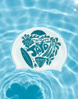 A white swim cap with a teal graphic design featuring the word 'JAYD' and tropical motifs like palm trees, a surfboard, and flowers, floating on rippling blue pool water.