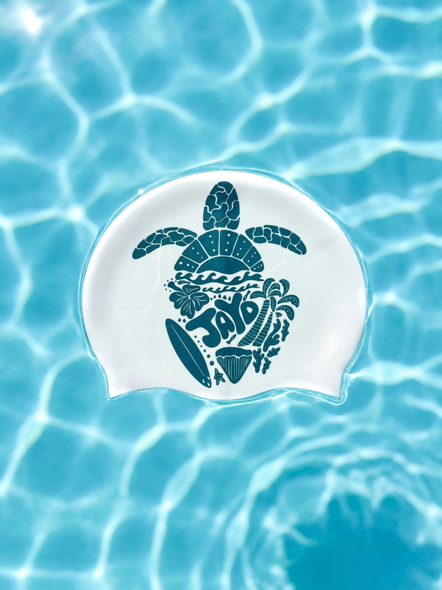 A white swim cap with a dark teal sea turtle graphic and the word 'JAYD' in a wave pattern, floating on clear blue water with ripples and light reflections.