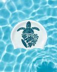 A white swim cap with a dark teal sea turtle graphic and the word 'JAYD' in a wave pattern, floating on clear blue water with ripples and light reflections.