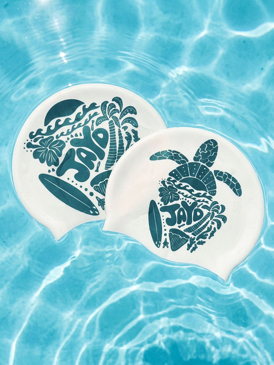 Two white swim caps with dark teal tropical and ocean-themed designs, floating on clear blue pool water.