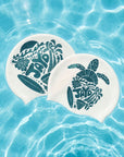 Two white swim caps with dark teal tropical and ocean-themed designs, floating on clear blue pool water.