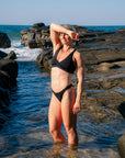 JAYD model Dom wearing the black Vienna bikini top and Amalfi bottoms as she stands in shallow ocean water on a rocky shoreline, with waves and rocks behind her under a clear blue sky.