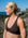 JAYD model in profile wearing the Cacao Vienna bikini top, standing outdoors on a rocky shoreline with the ocean and blue sky in the background.