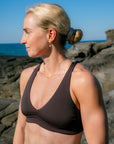 JAYD model in profile wearing the Cacao Vienna bikini top, standing outdoors on a rocky shoreline with the ocean and blue sky in the background.
